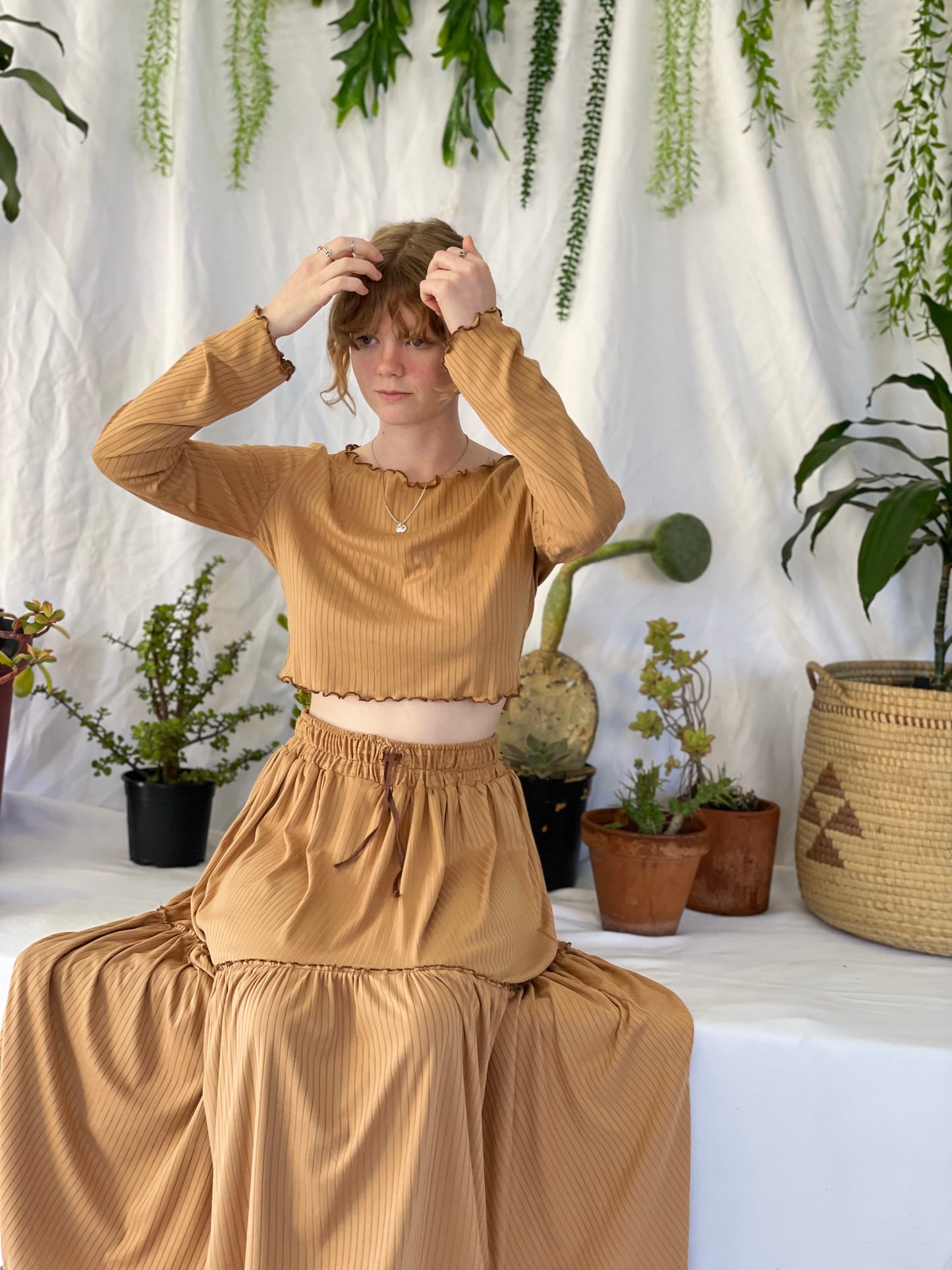 Model wears matching skirt and top set with silver necklace. The model sits in front of a white backdrop with pot plants.