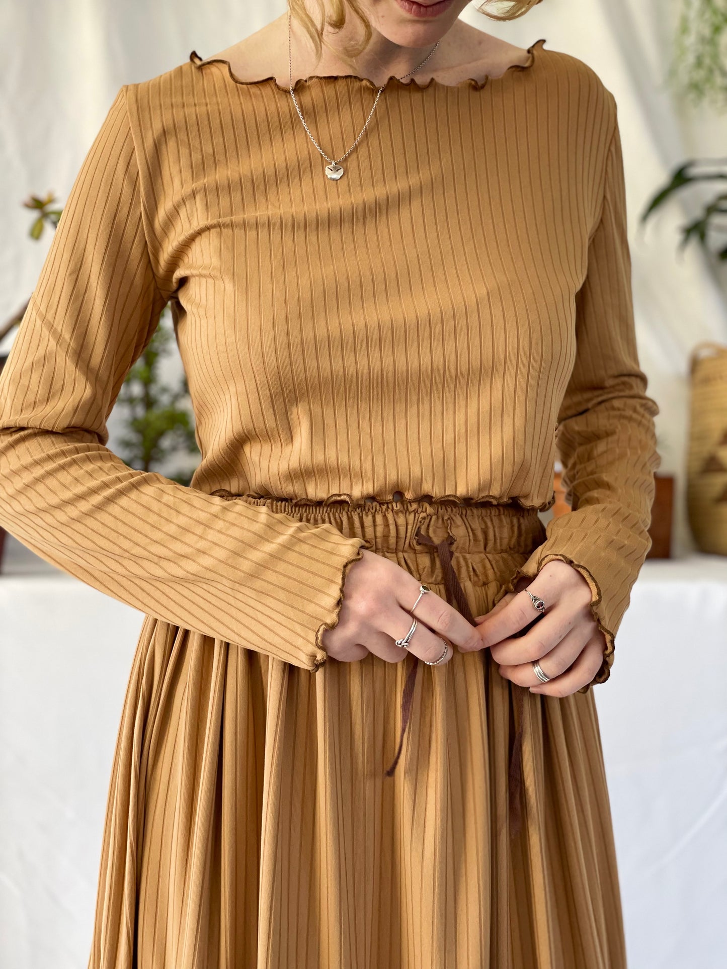 Model wears matching skirt and top set with a silver necklace. The model stands in front of a white backdrop with pot plants.