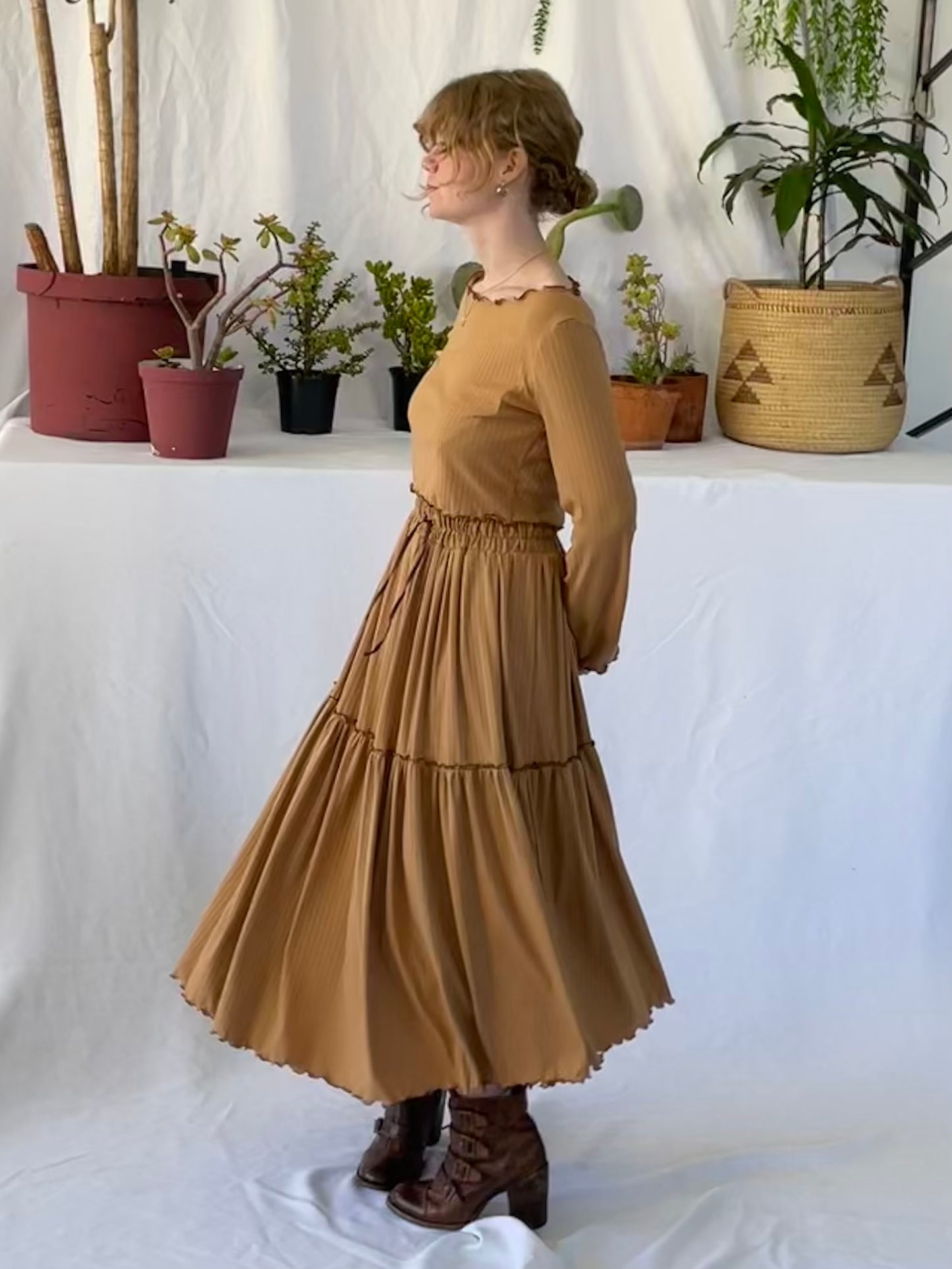 Model wears matching skirt and top set with brown boots. The model stands in front of a white backdrop with pot plants.