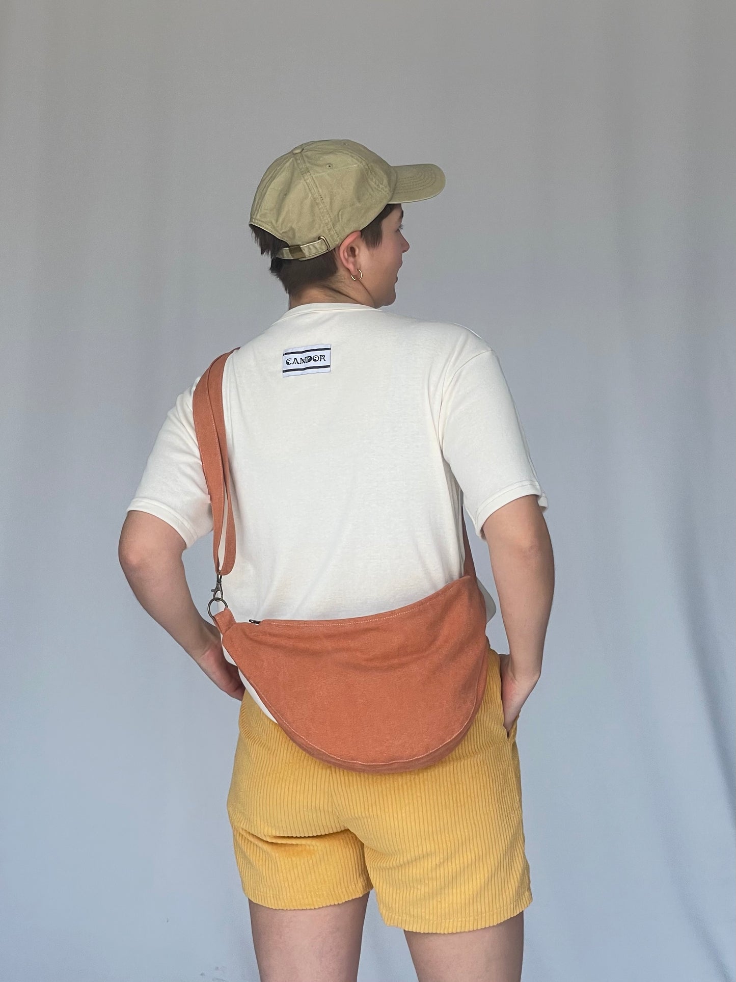 Models wears orange sling bag across body with a cream t-shirt, cap and yellow corduroy shorts against a white backdrop