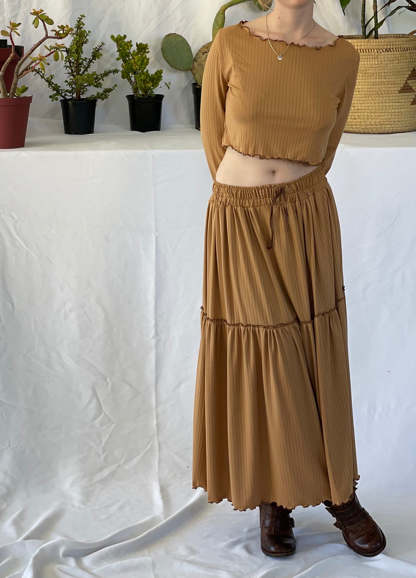Model wears matching skirt and top set with brown boots. The model stands in front of a white backdrop with pot plants.