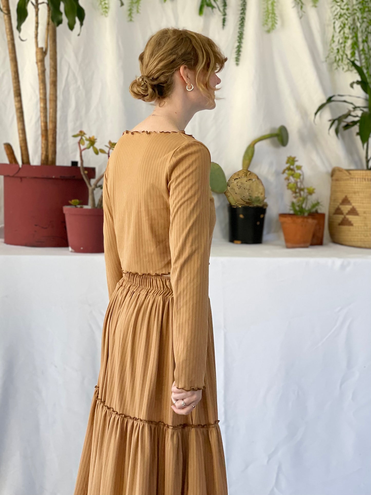 Model wears matching skirt and top set with her back to the camera. The model stands in front of a white backdrop with pot plants.