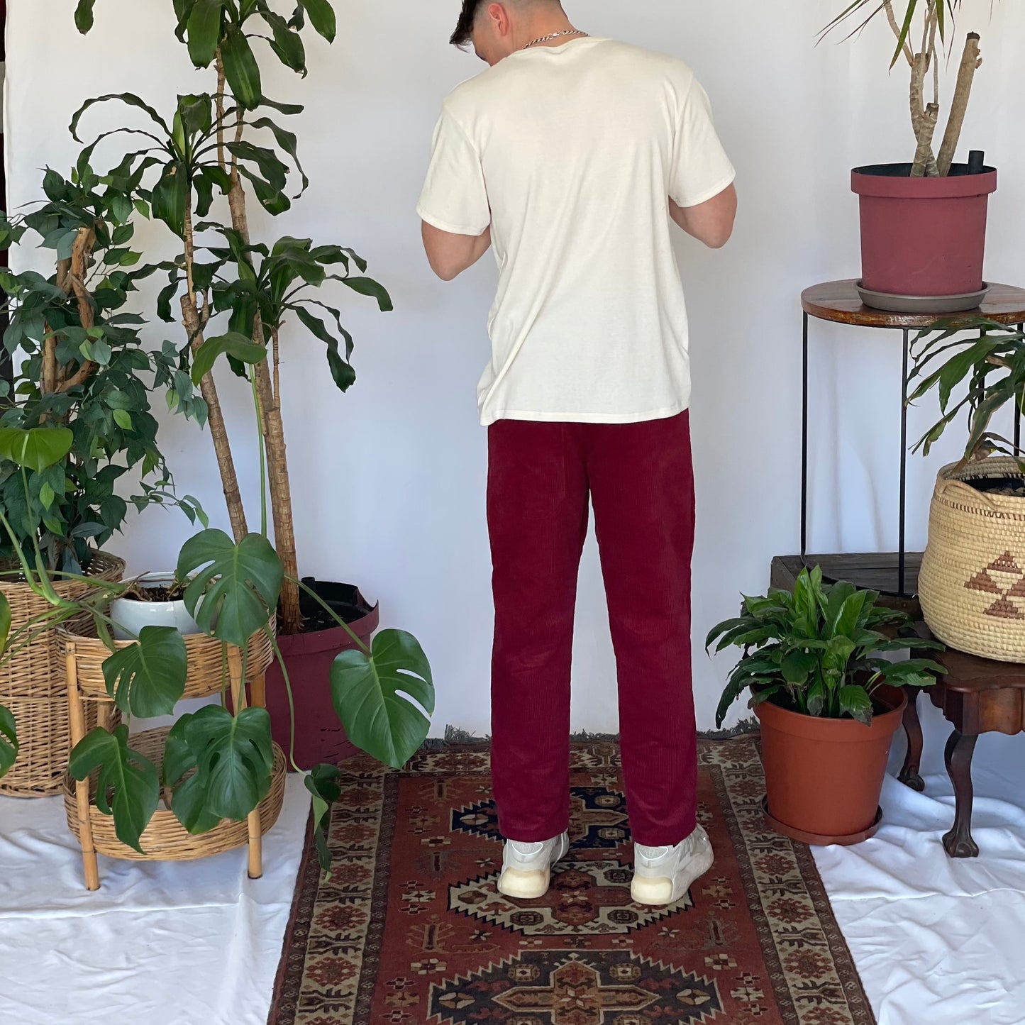 Model wears a cream t-shirt with burgundy corduroy pants and sneakers with their back to the camera. The model is standing on a narrow carpet against a white backdrop with plants and hanging foliage.