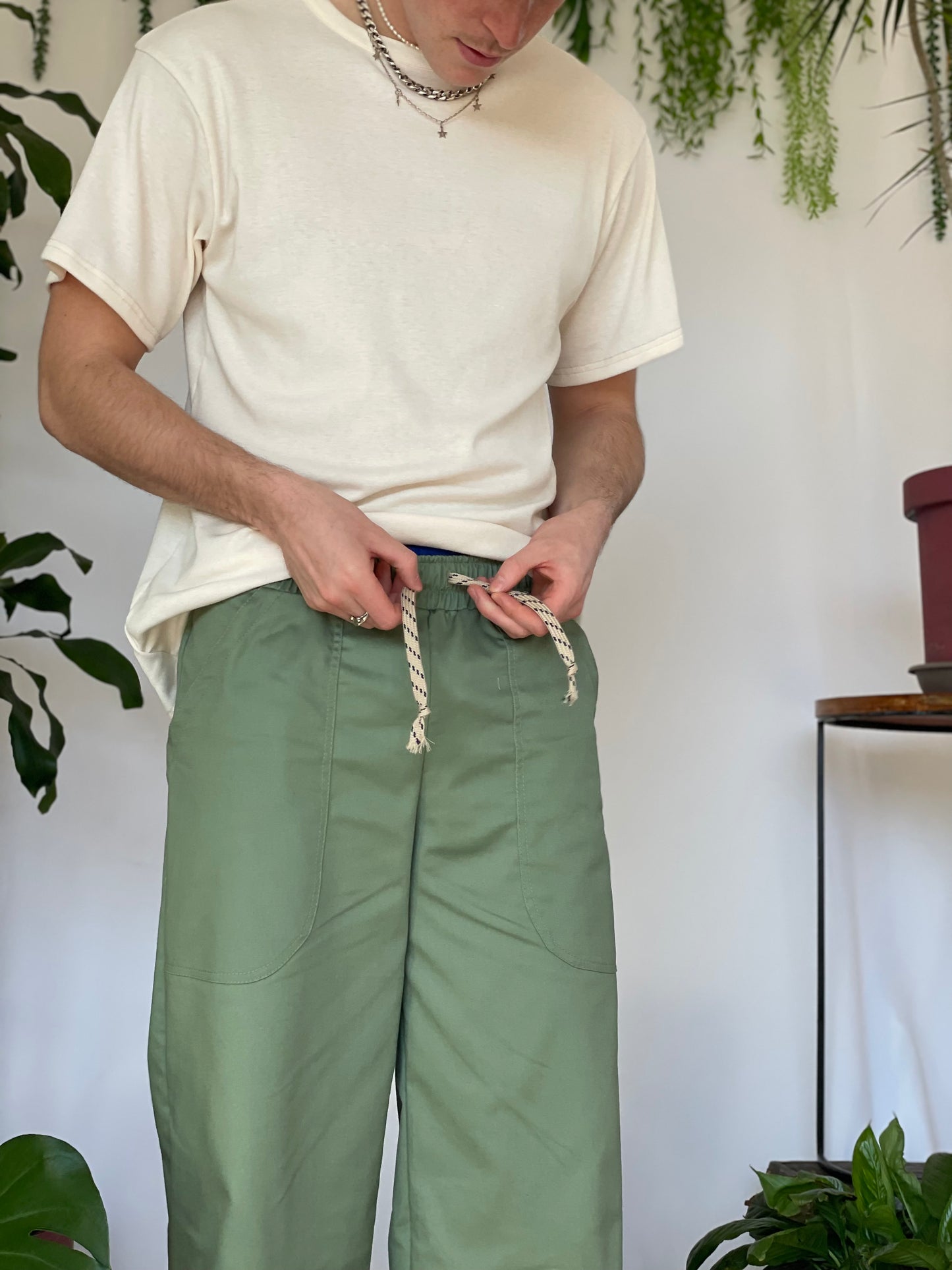 Model wears green pants with a cream t-shirt and silver jewellery. Model stands against a white backdrop with plants and hanging foliage.