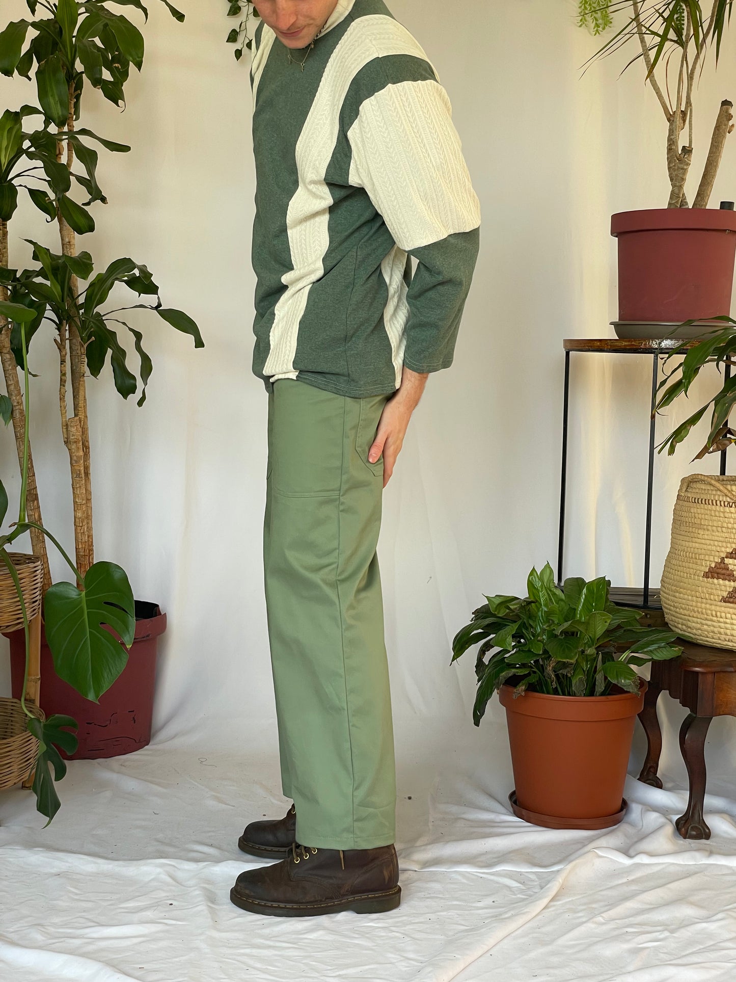 Model wears green pants with a striped jersey and green beanie. Model stands against a white backdrop with plants and hanging foliage.