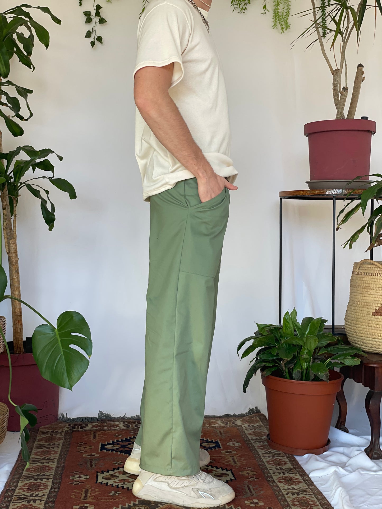 Model wears green pants with a cream t-shirt and silver jewellery. Model stands against a white backdrop with plants and hanging foliage.