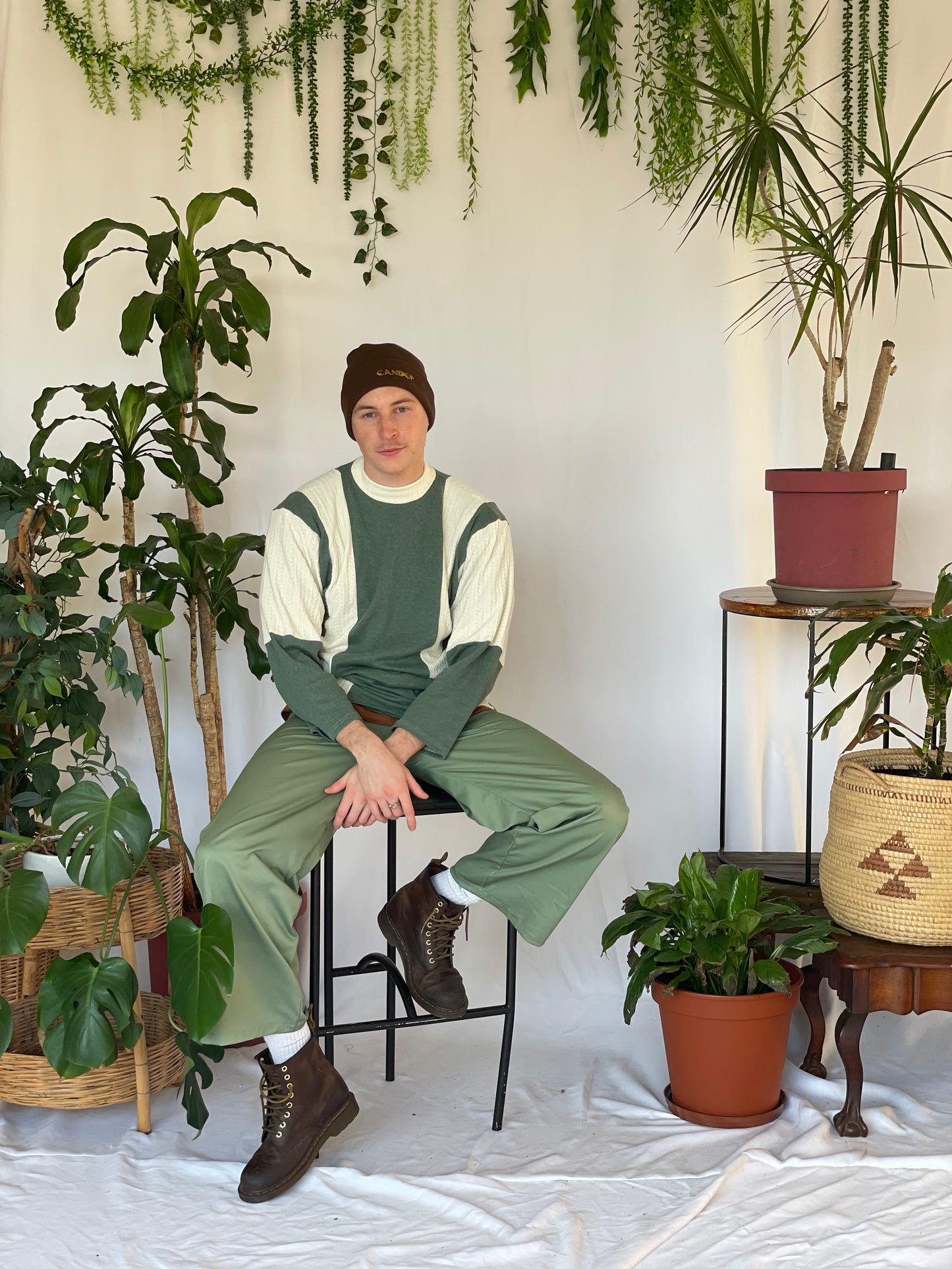 Model wears green pants with a striped jersey and green beanie. Model sits against a white backdrop with plants and hanging foliage.