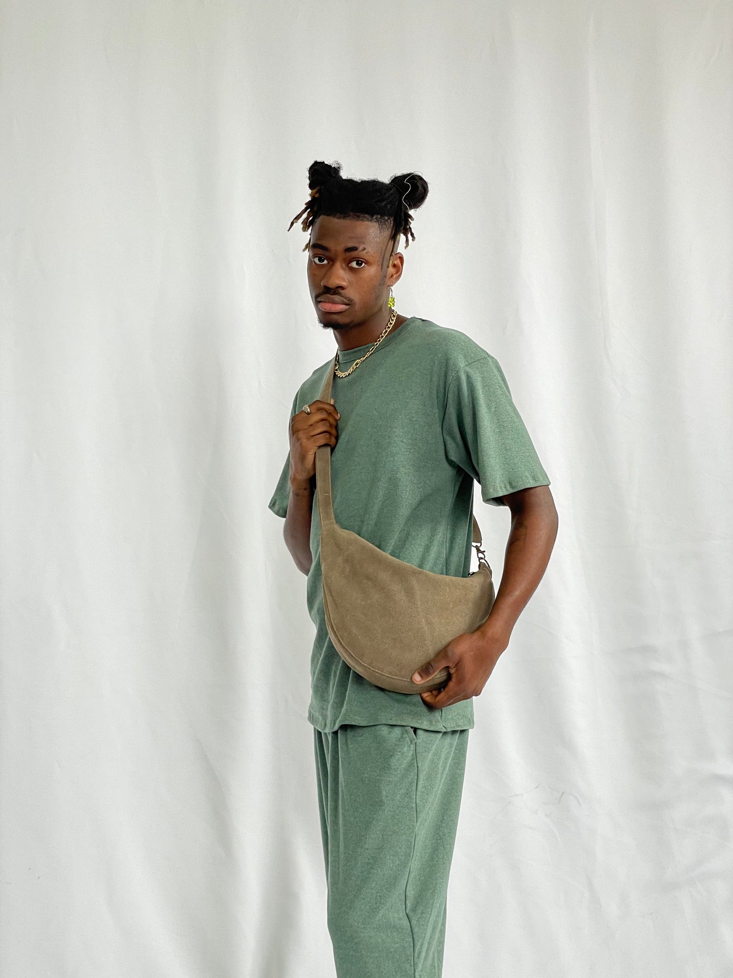 Model wears brown sling bag with green t-shirt and pants against a white backdrop
