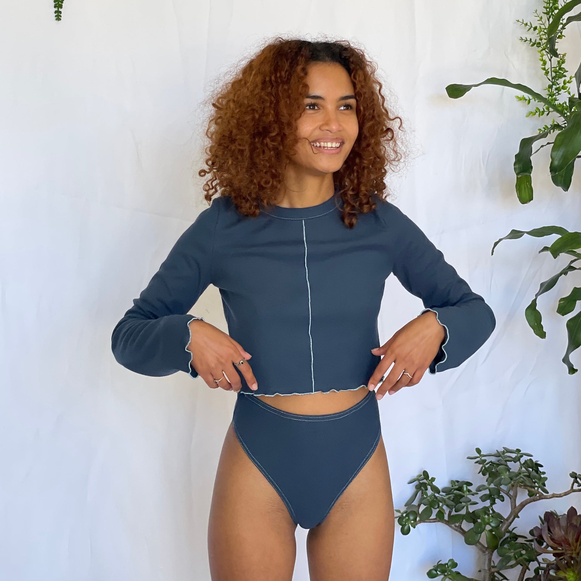 Model wears a navy blue underwear and long sleeve top on a white back drop with foliage. 