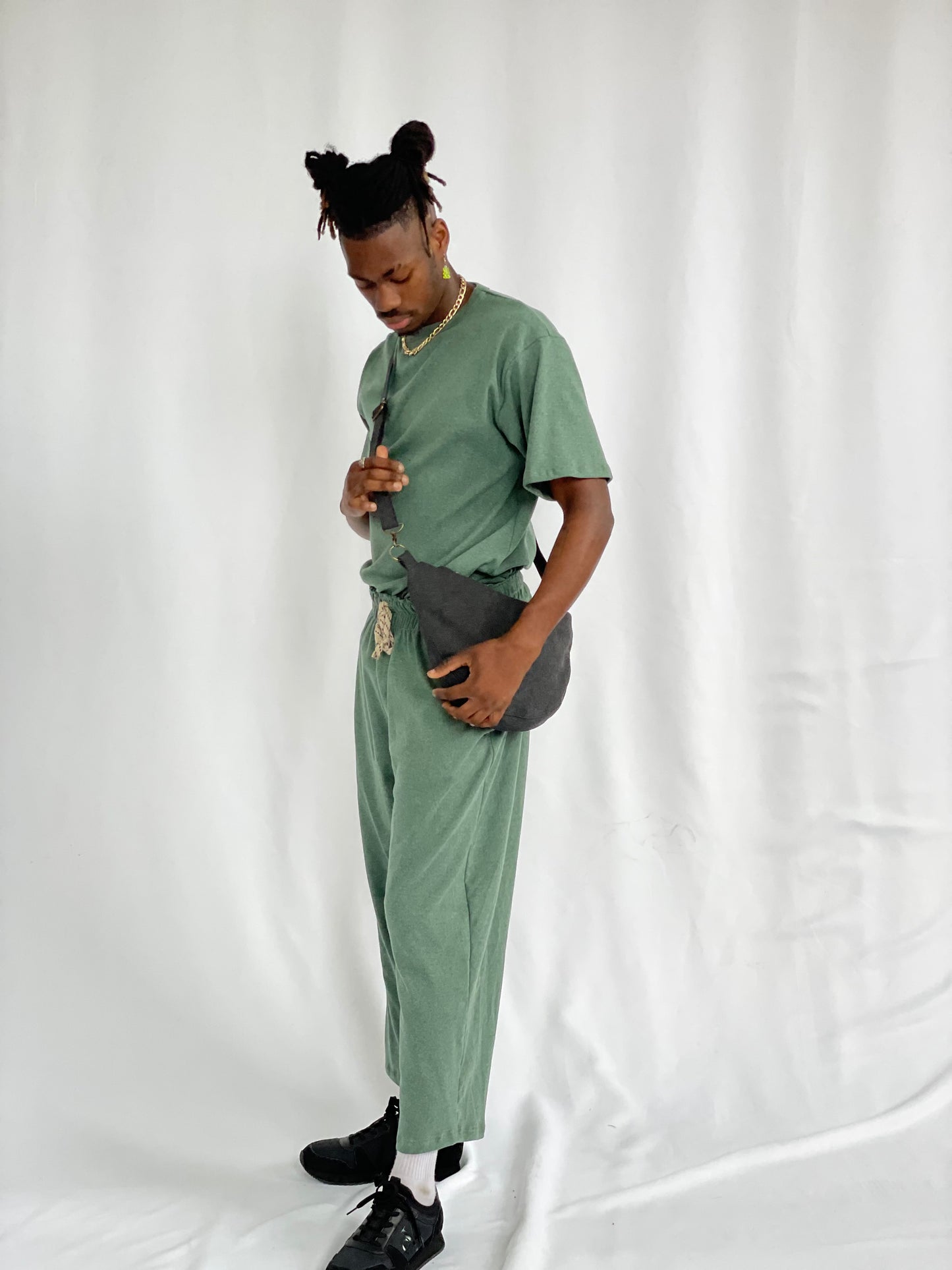 Model wears black sling bag cross body with green pants and t-shirt against a white backdrop 