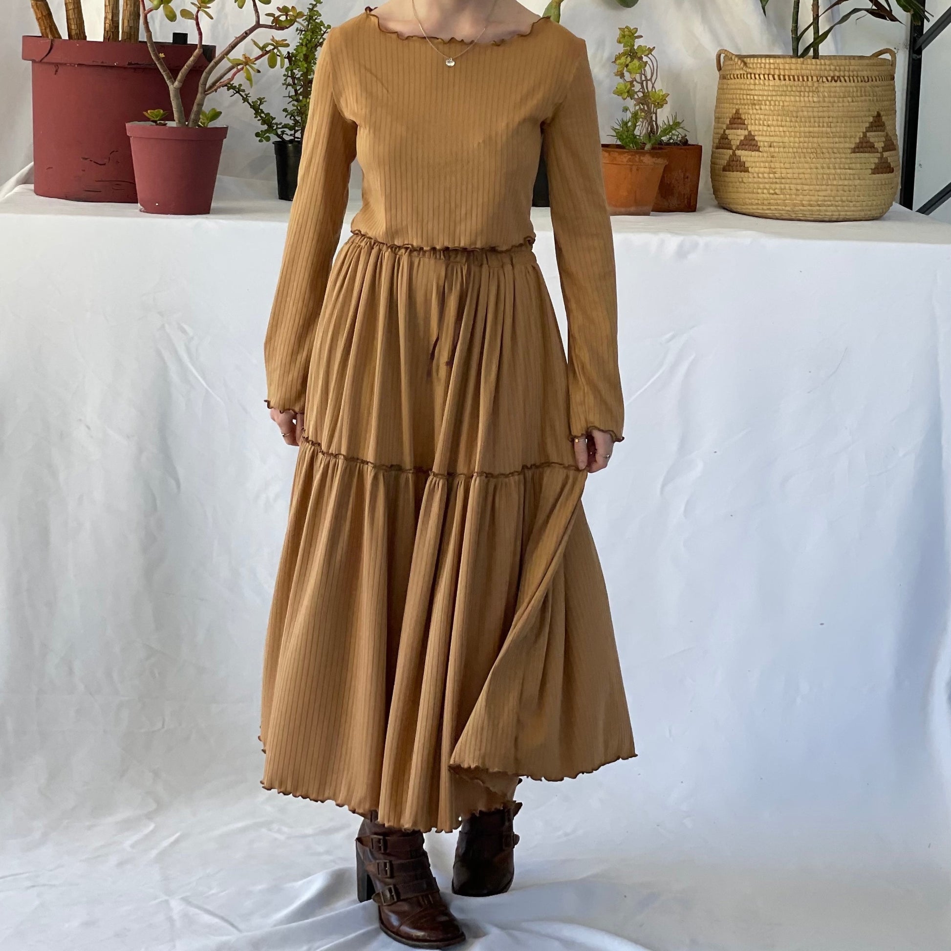 Model wears matching skirt and top set with brown boots. The model stands in front of a white backdrop with pot plants. 