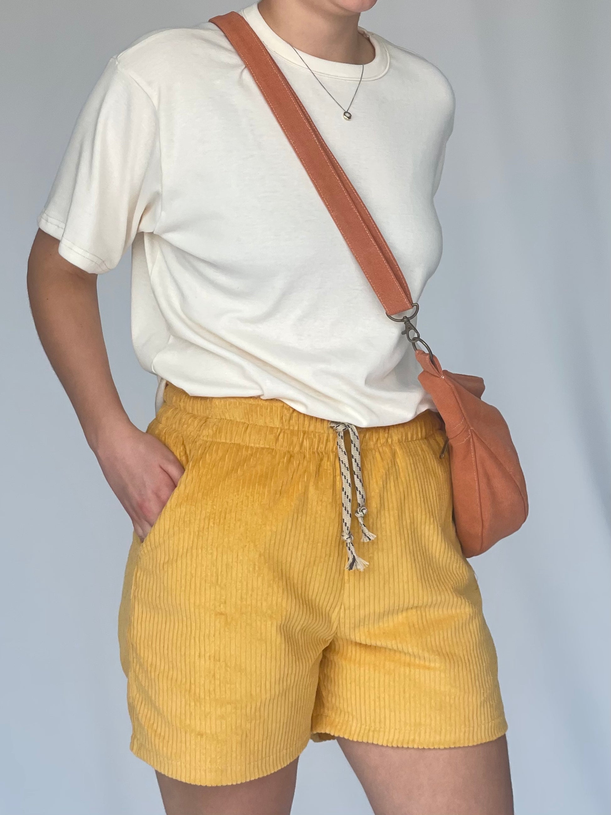Models wears orange sling bag across body with cream t-shirt and yellow corduroy shorts against a white backdrop.