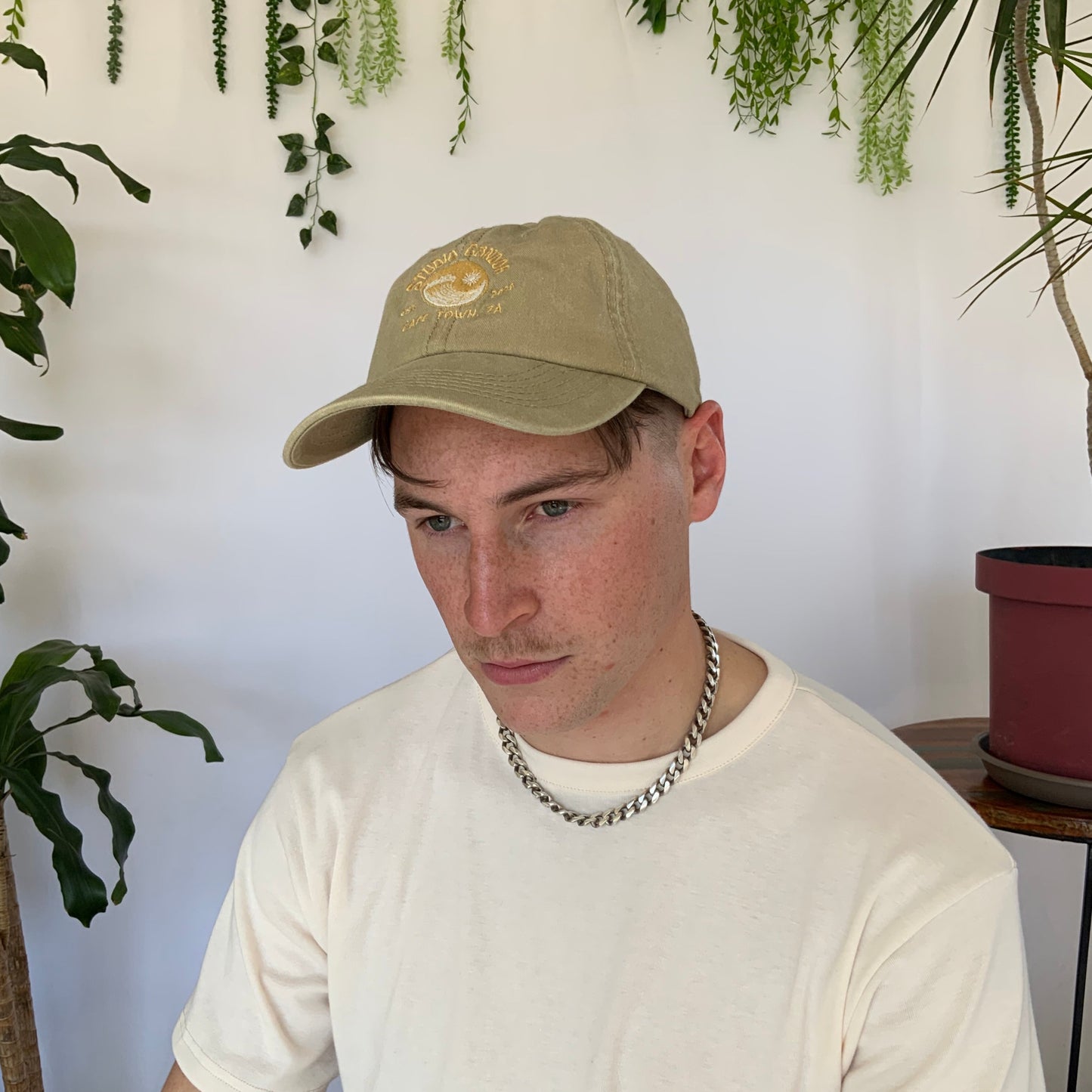 Model wears a stone coloured peak cap with a cream t-shirt and silver necklace. Model sits against a white backdrop with plants and hanging foliage.