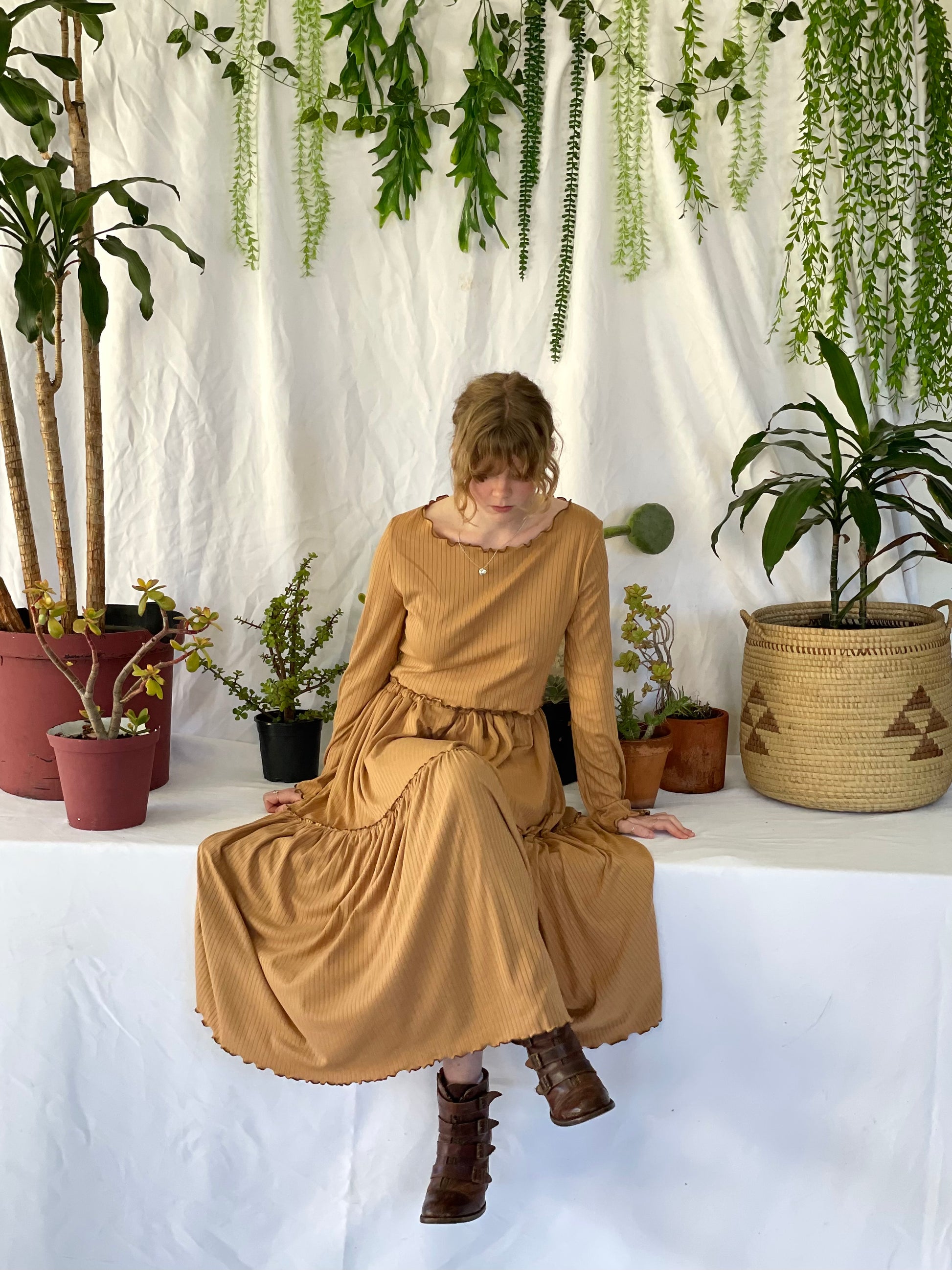 Model wears matching skirt and top set with brown boots. The model stands in front of a white backdrop with pot plants. 