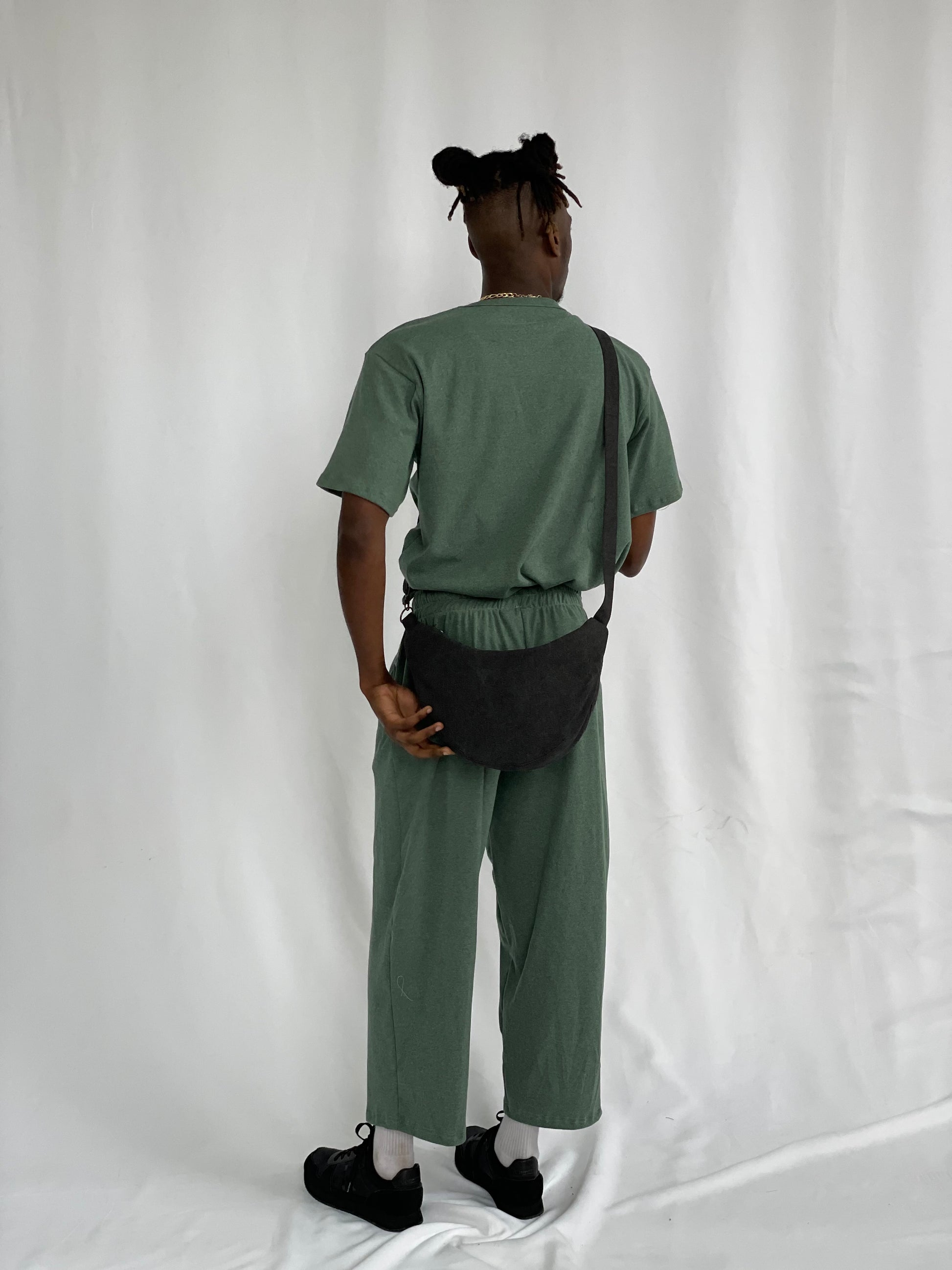 Model wears black sling bag with green t-shirt and pants against a white backdrop.