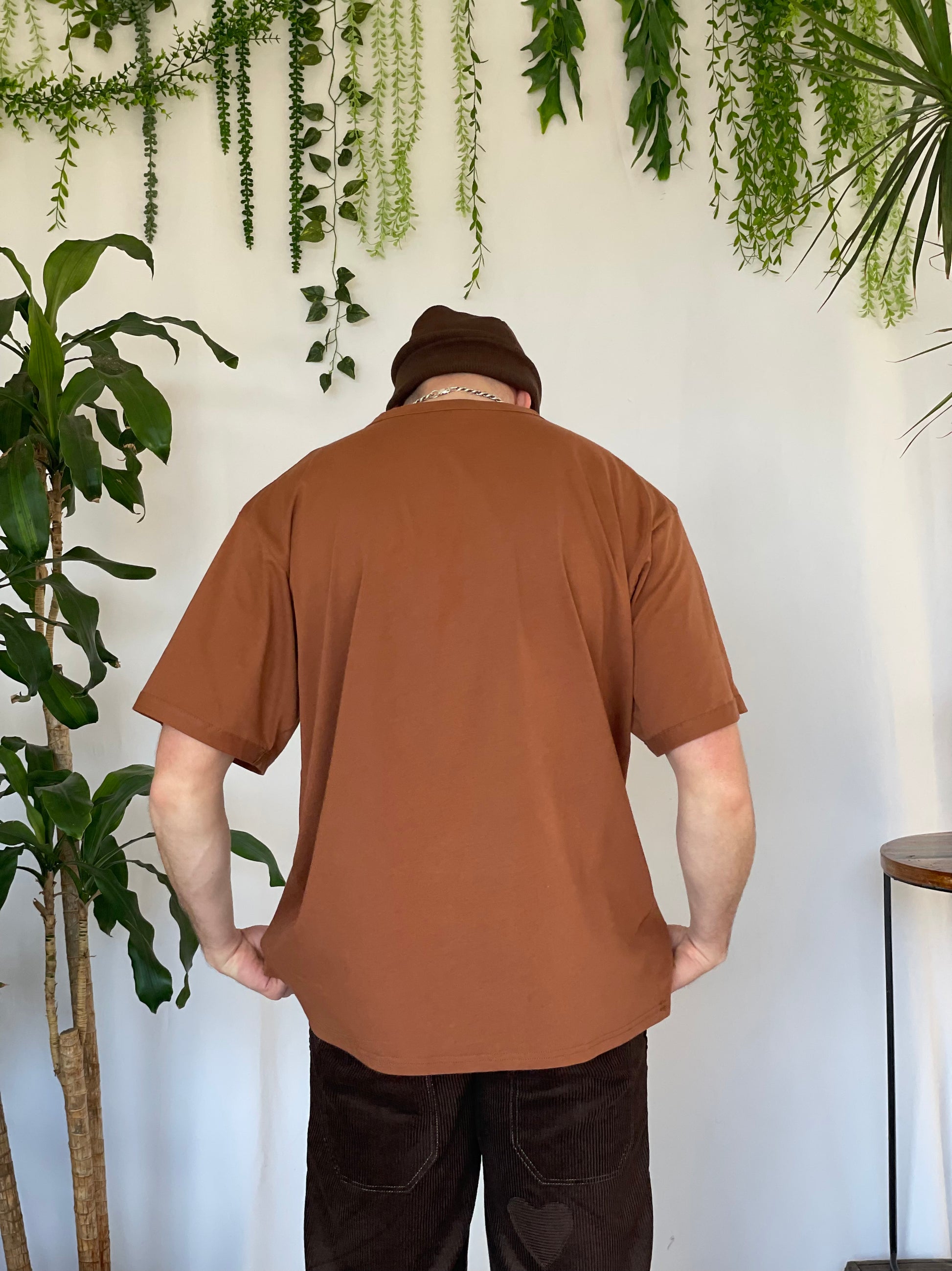 Model stands with back to camera wearing a light brown t-shirt, brown corduroy pants and brown beanie against a white backdrop with plants and hanging foliage.