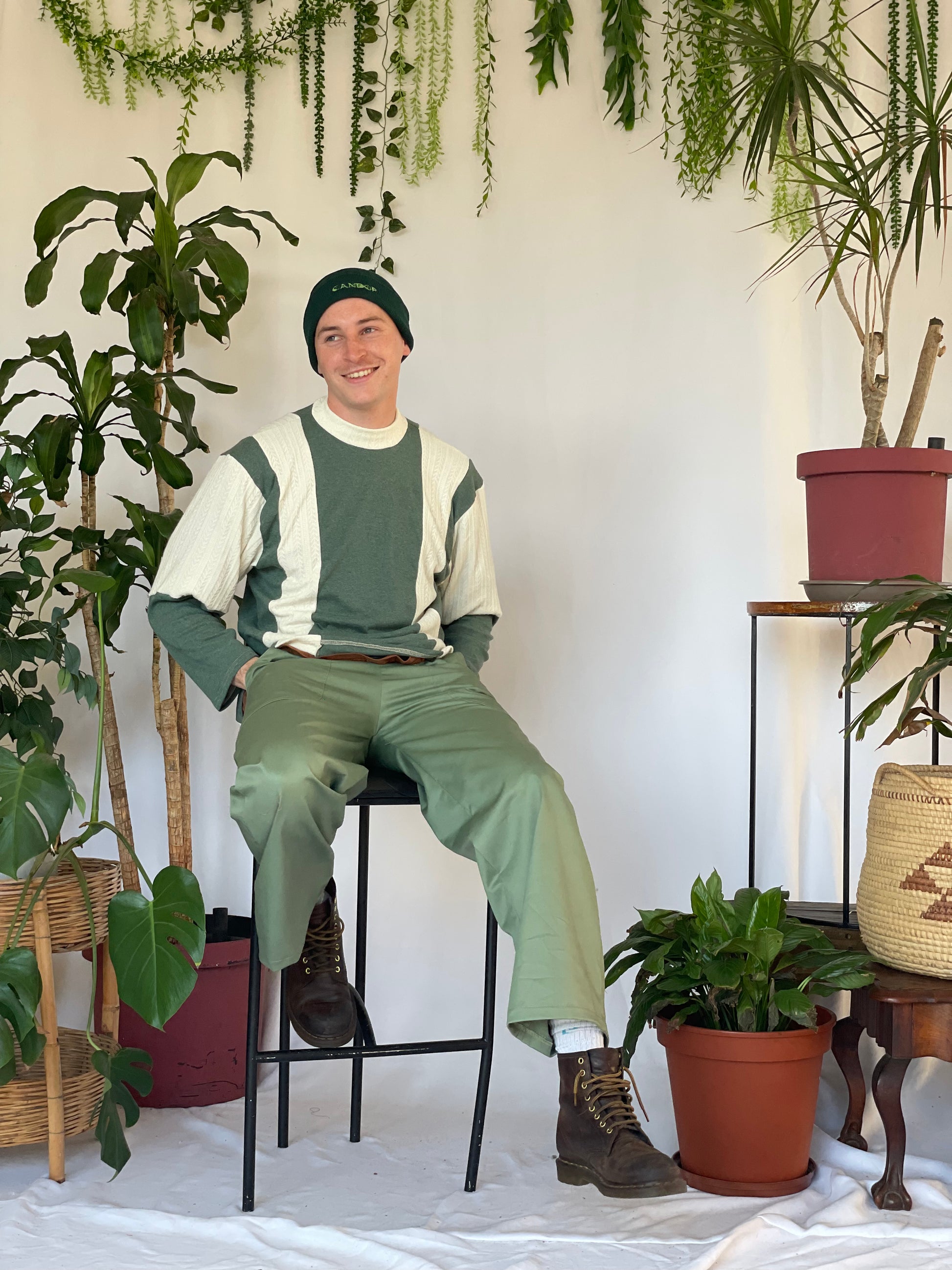 Model wears green pants with a striped jersey and green beanie. Model sits against a white backdrop with plants and hanging foliage.