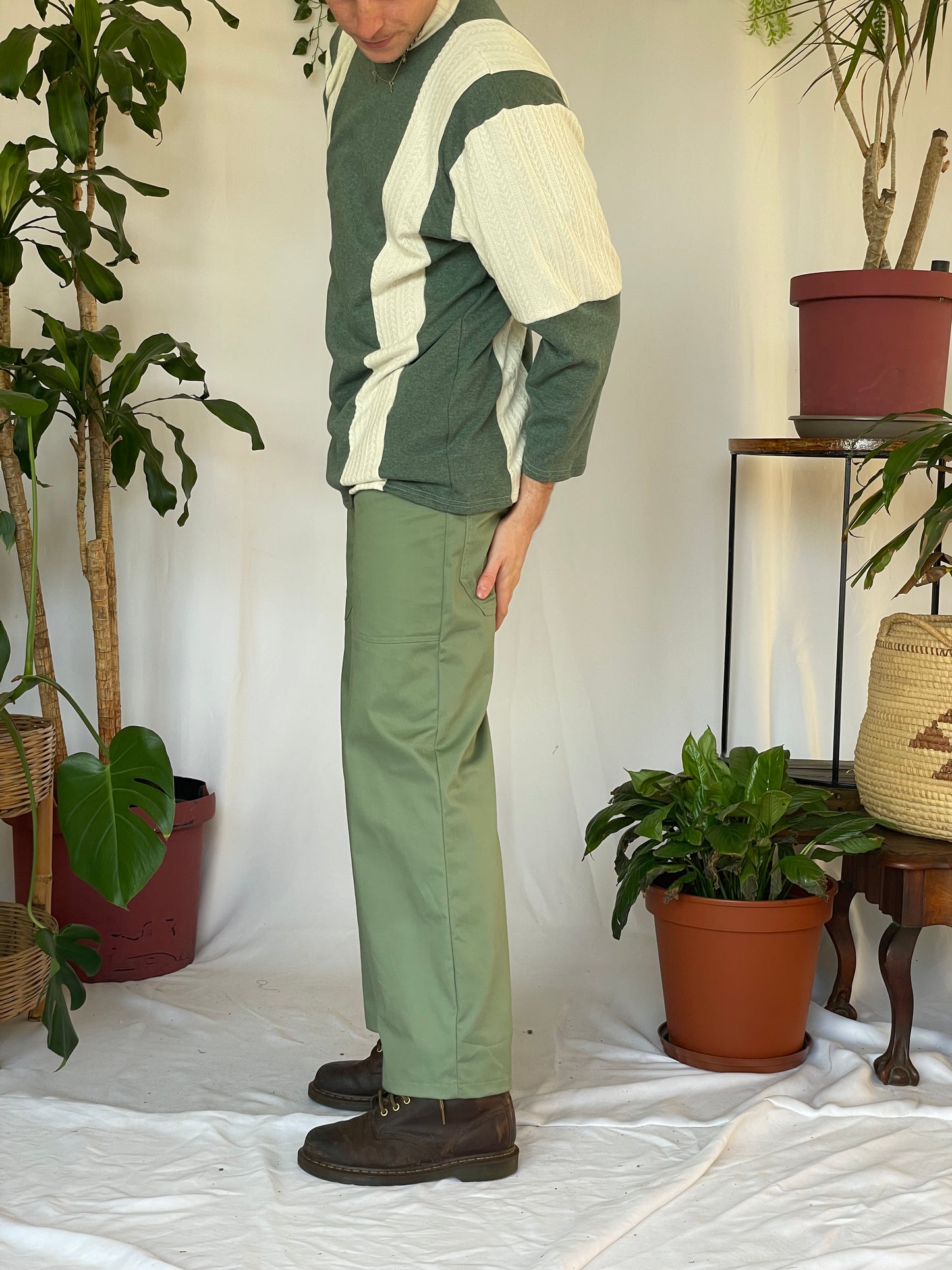 Model wears green pants with a striped jersey and green beanie. Model stands against a white backdrop with plants and hanging foliage. 