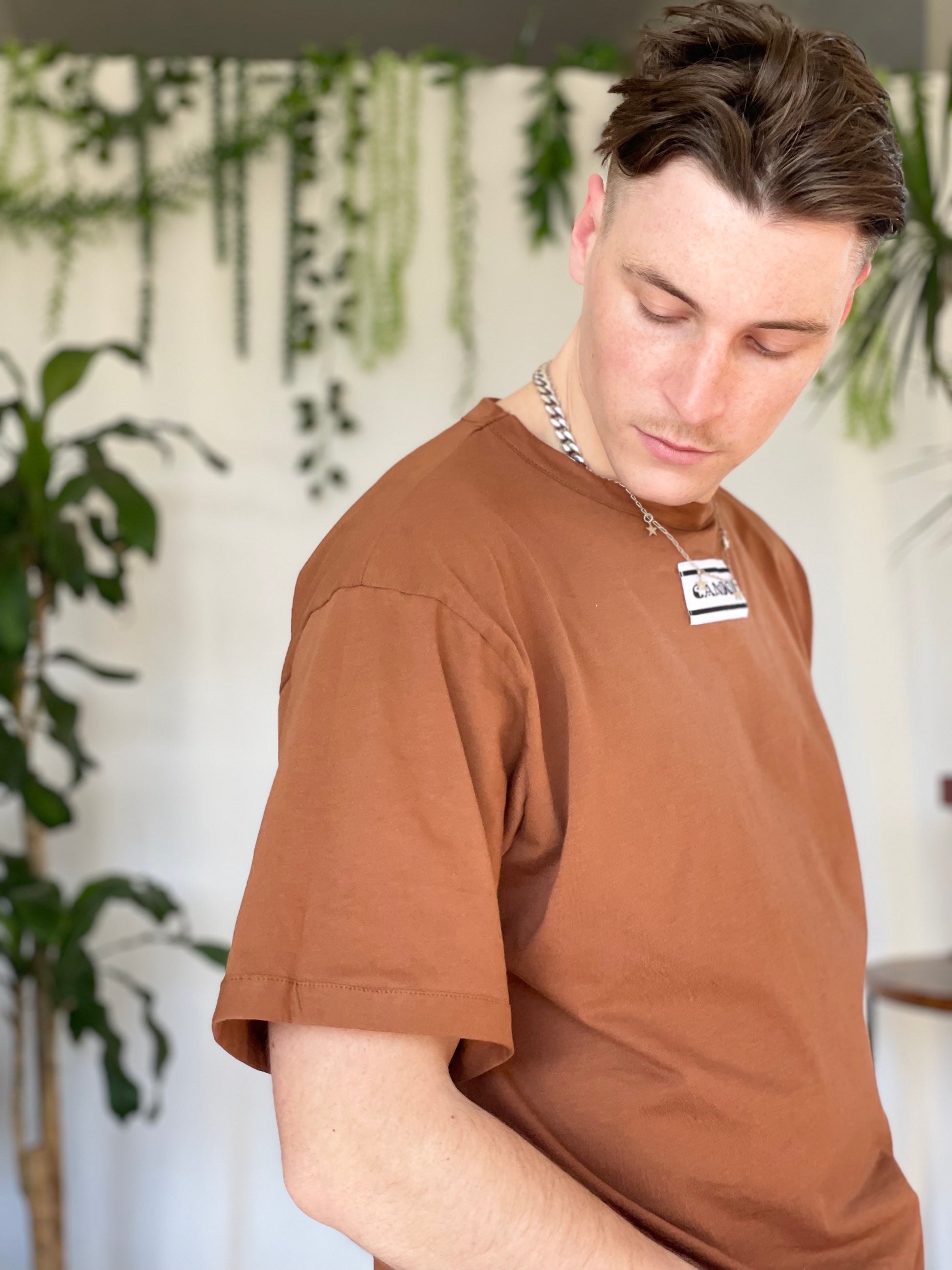 Models wears light brown t-shirt with Candor label. Model stands side on looking down against a white backdrop with plants.
