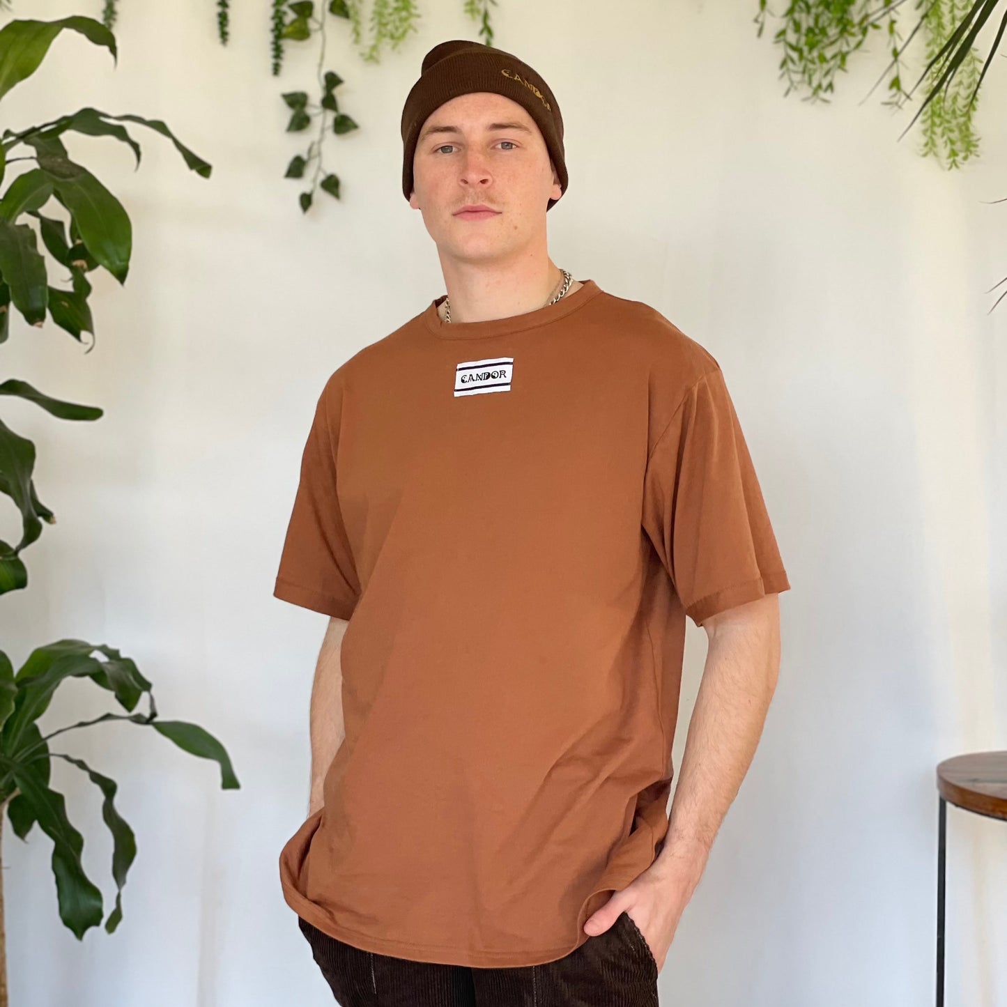 Models wears light brown t-shirt with Candor label, brown Candor beanie and brown corduroy pants with hands in pockets. Model stands against a white backdrop with plants and hanging foliage.