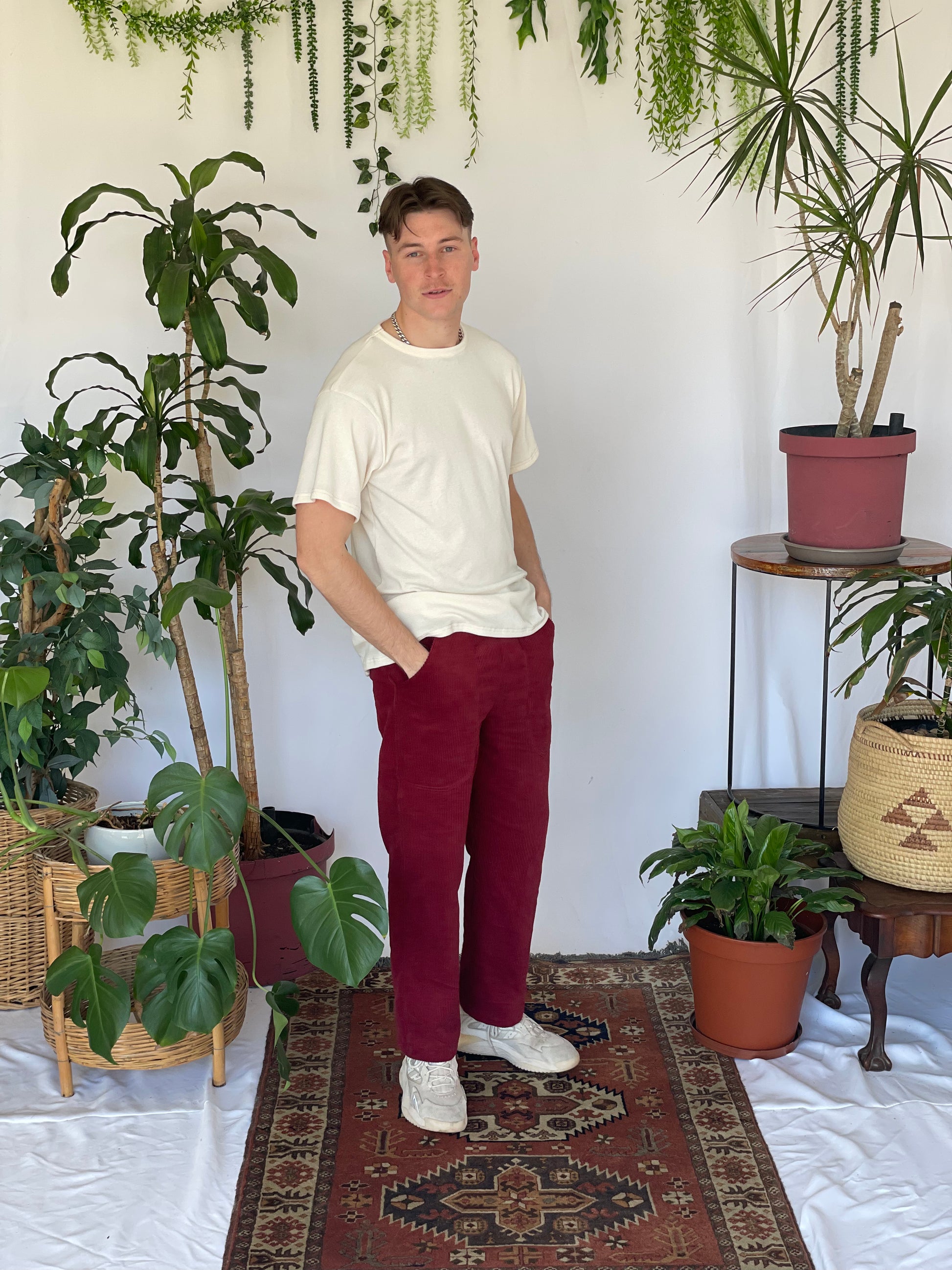 Model wears a cream t-shirt with burgundy corduroy pants and sneakers with their hands in pockets. The model is standing on a narrow carpet against a white backdrop with plants and hanging foliage. 