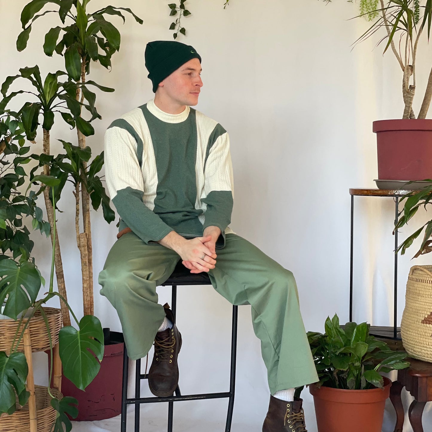 Model wears green pants with a striped jersey and green beanie. Model sits against a white backdrop with plants and hanging foliage.