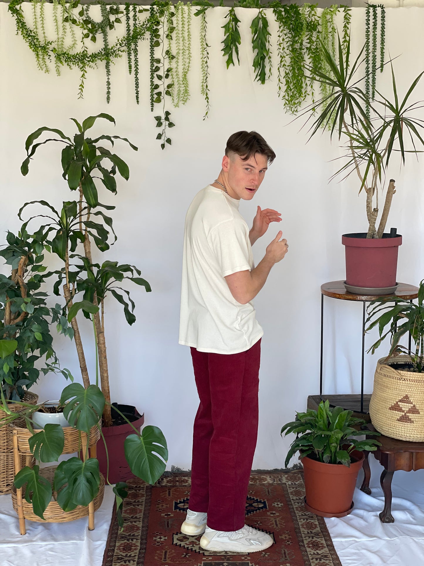 Model wears a cream t-shirt with burgundy corduroy pants and sneakers side on to the camera. The model is standing on a narrow carpet against a white backdrop with plants and hanging foliage.
