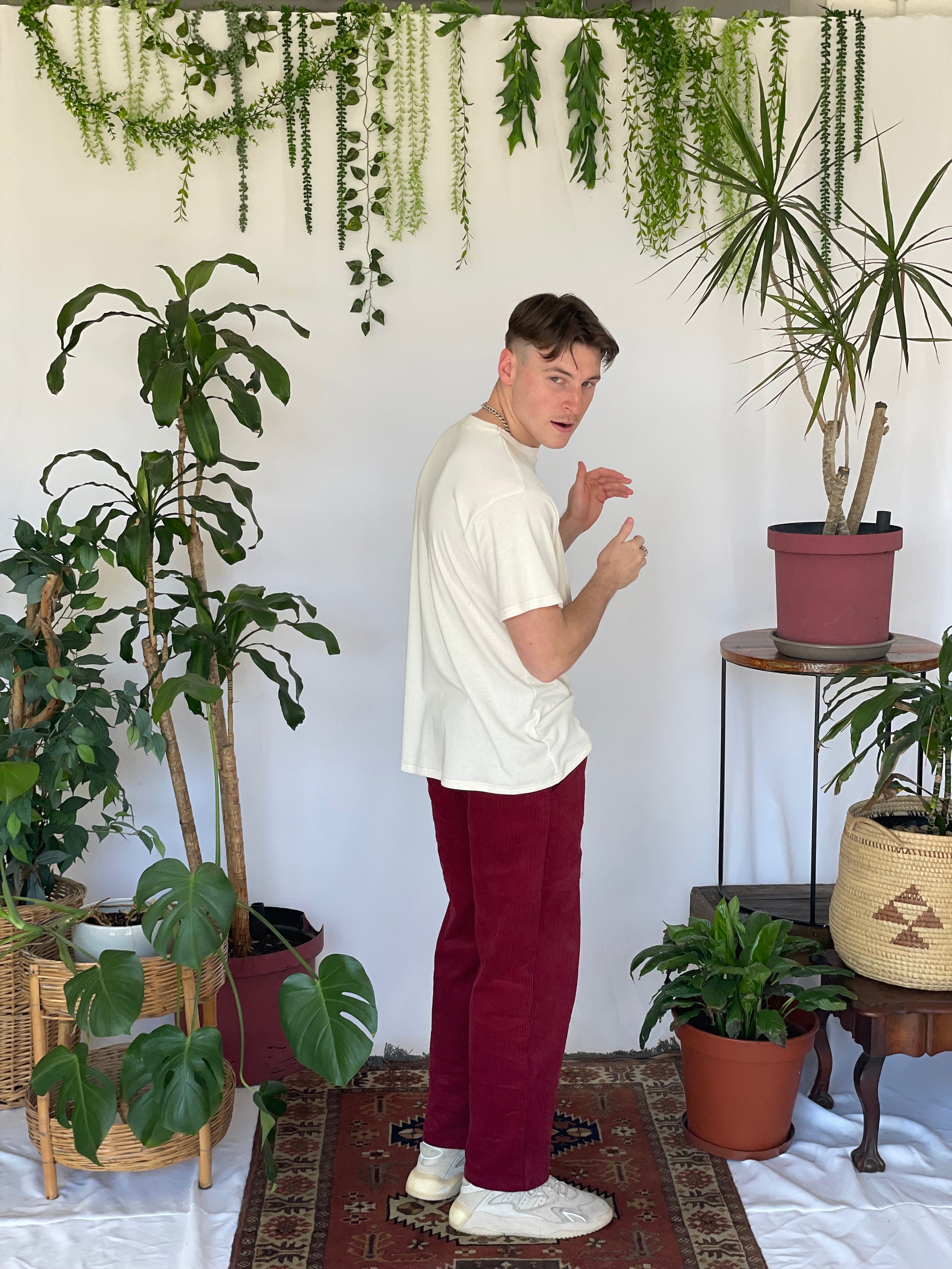 Model wears a cream t-shirt with burgundy corduroy pants and sneakers side on to the camera. The model is standing on a narrow carpet against a white backdrop with plants and hanging foliage.